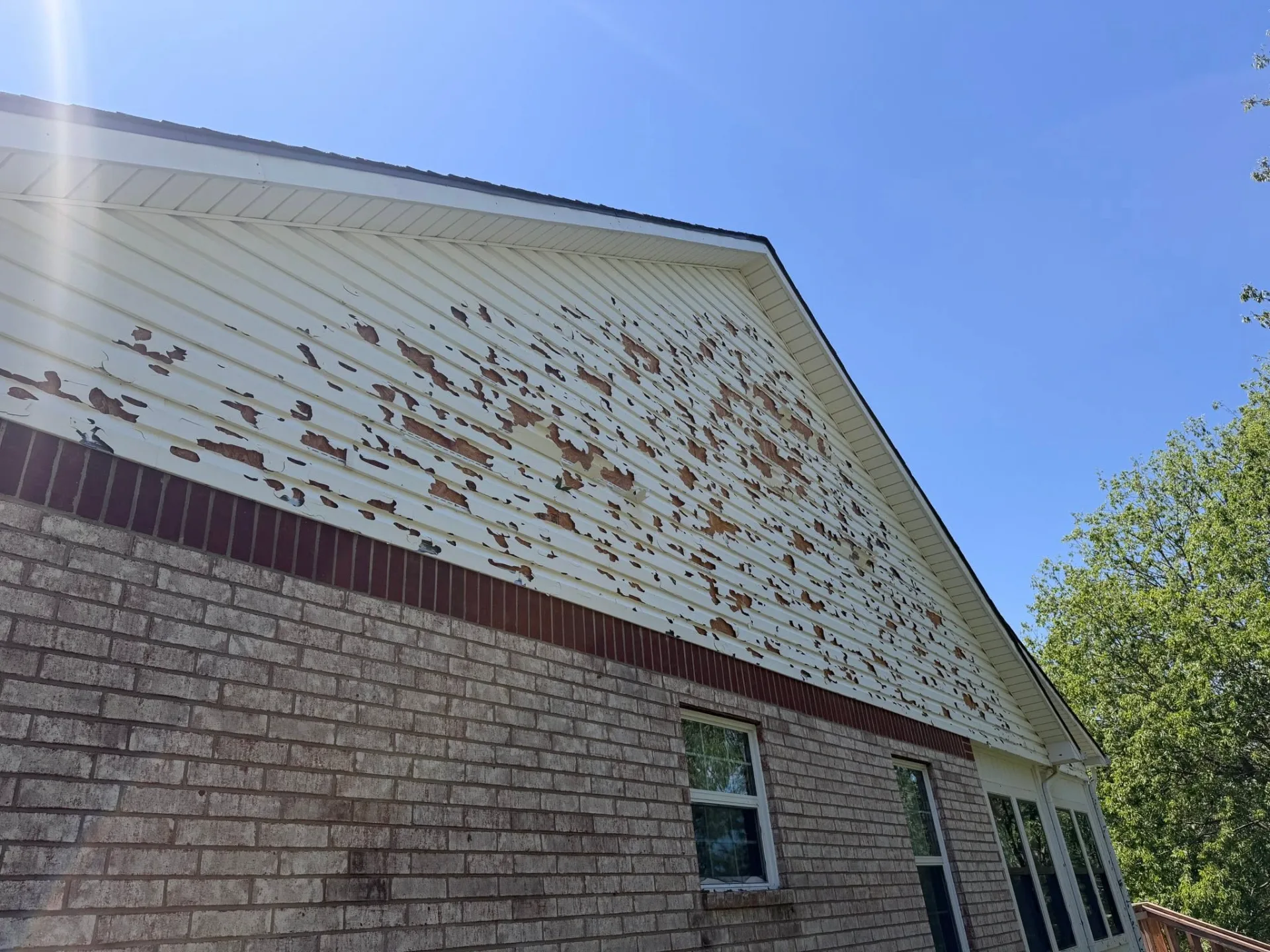 Hail-damaged white vinyl siding with widespread paint chipping on a Claremore home over brick